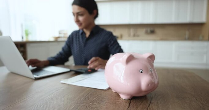 Close up focus on pink piggy bank on table, on background Indian housewife calculate monthly expenses, manage budget, pay utility bills via e-bank use laptop. Save money for future, finances analysis