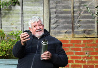 Senior man sitting outside, smiling with a flask and a drink in his hand.