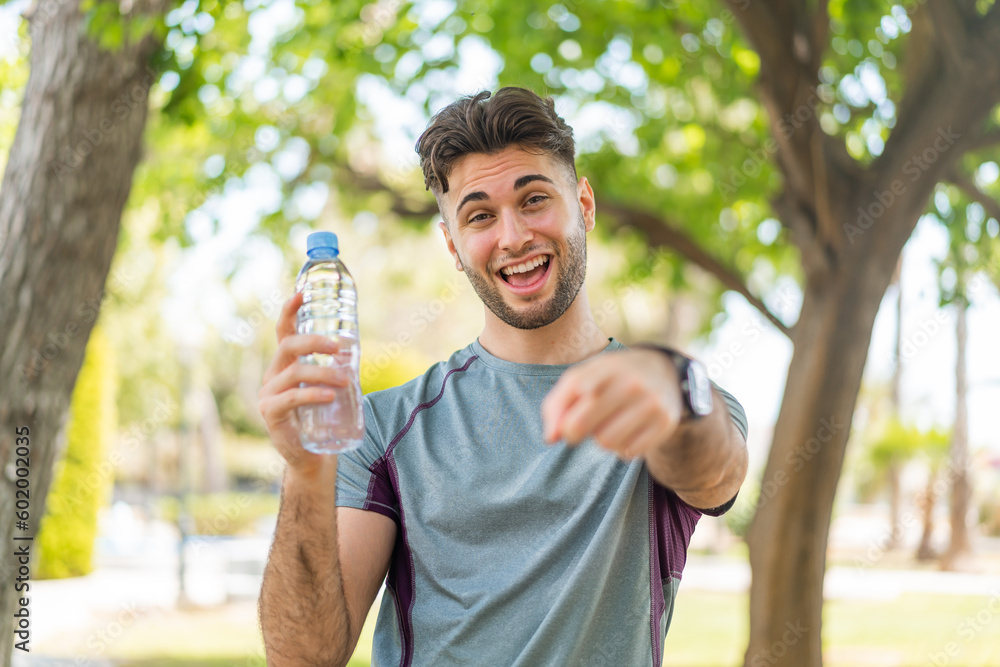 Wall mural Young handsome sport man with a bottle of water at outdoors points finger at you with a confident expression