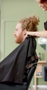 Vertical video of caucasian female hairdresser putting cape on client at hair salon, in slow motion
