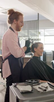 Vertical video of caucasian male hairdresser dyeing client's hair at hair salon, in slow motion