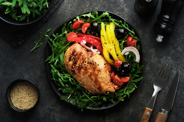Salad with grilled chicken breas fillet with fresh arugula, tomatoes and olives on black plate on old dark background. Chicken meat with salad. Ketogenic lunch. Healthy food. Top view.