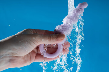 Woman holding lilac anal beads under running water on blue background. Sex toy hygiene concept.