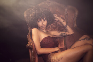 Tattoed alternative young woman with natural afro hair and piercings posing on a victorian armchair...