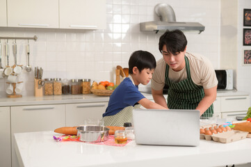 Happy young Asian father and kid are using a laptop computer to learn how to cooking food in their home kitchen. - obrazy, fototapety, plakaty
