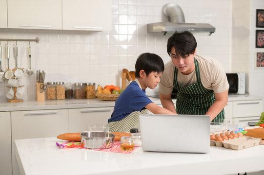 Happy young Asian father and kid are using a laptop computer to learn how to cooking food in their home kitchen.
