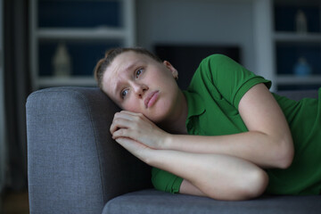 Worried young woman sit on sofa look aside lean forward with folded hands feel fear anxiety. Teen...