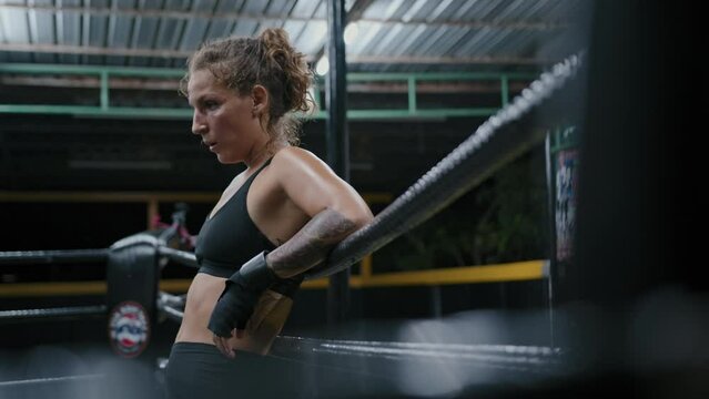 Woman muay thai boxer hanging on ring ropes resting between rounds. Tired female fighter wet with sweat breathes deeply during workout at gym