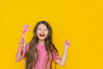 Portrait of a beautiful cheerful model of a girl with a big candy on a yellow background, a child with a lollipop on an isolated background, a glamorous model with a candy on a stick.