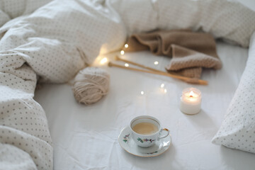 Cozy morning still life with a coffee cup, candle, needles and a woolen yarn in white bed. Knitting warm woolen sweater or scarf top view. Hygge lifestyle, cozy mood. Handicraft day concept. 