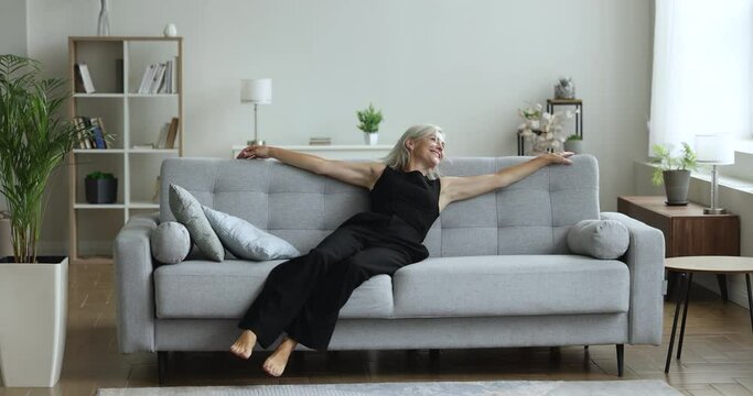 Cheerful active mature woman falling sofa in living room, resting on couch with open arms, enjoying relaxation, leisure at home, weekend, new comfortable furniture