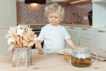 Little cute boy is having breakfast at the kitchen table. He is wearing a white T-shirt and has blond curly hair.