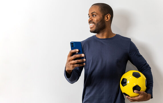 A dark-skinned man posing happily on a white background in casual clothes with a soccer ball and a mobile. The boy looks to the side, promoting the game, ad, football advertising.