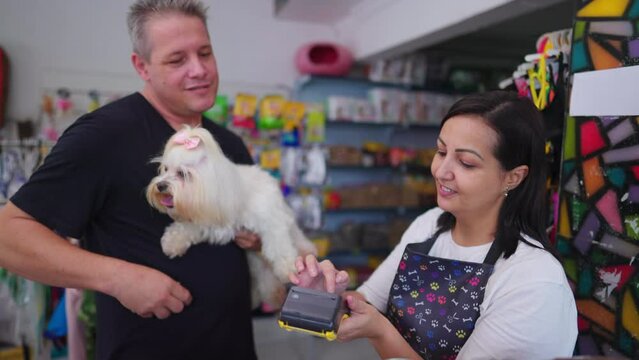 Customer paying for service with contactless credit card on payment machine standing inside Pet Shop holding small dog. Happy client pays for grooming procedure