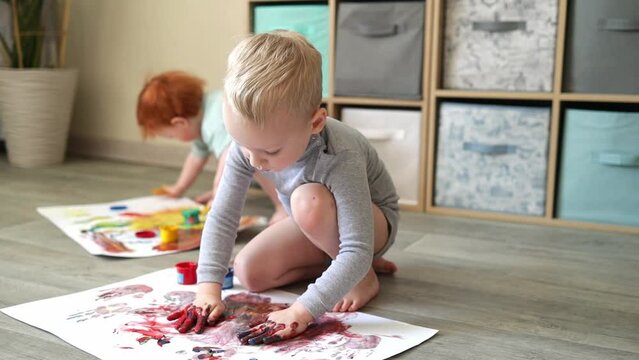 Small children in the home interior draw with paints on a sheet of paper with their own hands. Finger painting or art therapy for children