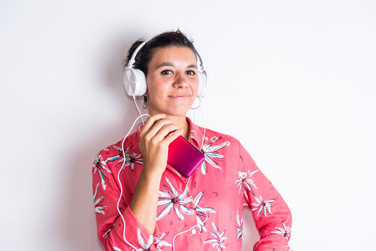 Attractive latina in flowered shirt listening to music on headphones from her cell phone, with neutral background, complicit look