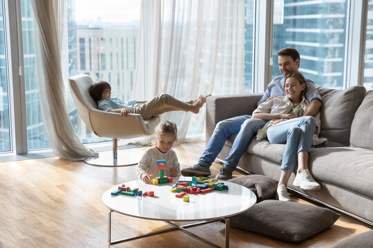 Couple of happy young parents resting on sofa in city apartment with large window, watching children, Two kids enjoying leisure at home, constructing toy building blocks, relaxing in armchair