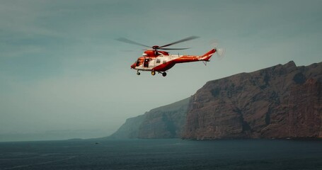 TENERIFE, CANARY ISLANDS, SPAIN, 2023 April: Rescue helicopter flies over a stormy ocean near rocky coastline. Los Gigantes cliffs.