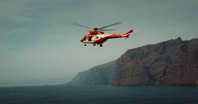 TENERIFE, CANARY ISLANDS, SPAIN, 2023 April: Rescue helicopter flies over a stormy ocean near rocky coastline. Los Gigantes cliffs.