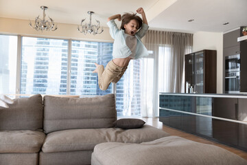 Excited crazy boy kid jumping on big sofa, throwing body on soft couch with wild scream, flying in...