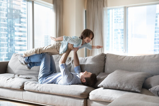 Strong dad lying on back on big soft couch in city apartment, lifting happy little son kid up with hands and legs support, keeping acroyoga balance, playing with child swimming in water,
