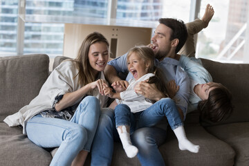 Happy young mom and dad tickling cheerful screaming little kids on sofa, laughing, having fun, playing with active playful children at home, enjoying family leisure, relationship - obrazy, fototapety, plakaty