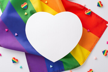 Top view flat lay of LGBT parade symbolic items, such as pin badges, heart-shaped trinkets, rainbow flag on a plain white background with an empty heart shaped card for text or advertisement