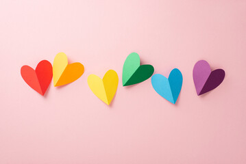 A flat lay top view of LGBT pride items: a rainbow colored paper hearts, placed on a pastel pink backdrop in line composition - obrazy, fototapety, plakaty