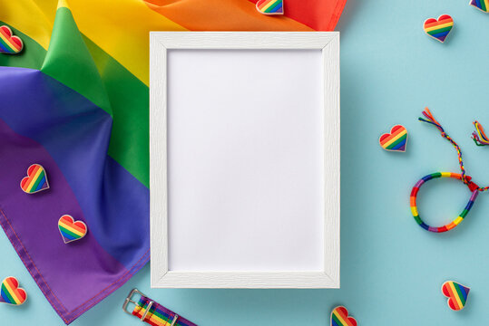 LGBT History Month essentials arranged in a top-down view. Rainbow flag, pin badges, colorful hearts, wristlet, laid out on a pastel blue background with an empty photo frame for message or picture