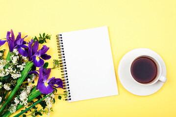 Coffee cup, blank notepad with pen and bouquet of blue iris flowers on yellow background. Top view, flat lay, mockup