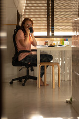 An adult woman drinking coffee at kitchen table - looking in the camera