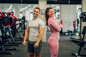 Fototapeta na wymiar The sporty happy muscular couple is posing in a gym while smiling at the camera.