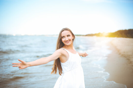 Portrait of a happy smiling woman in free happiness bliss on ocean beach standing with a hat and sunglasses. A female model in a white summer dress enjoying nature during travel holidays vacation