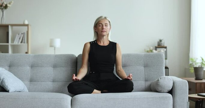 Calm peaceful mature woman with closed eyes meditating at home, sitting on soft couch, keeping eyes closed, lotus posture, zen hands, practicing, training mindfulness, concentration, yoga