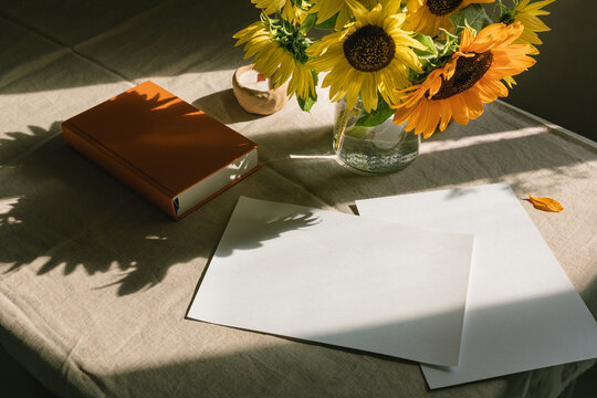 Bunch of sunflowers in a vase next to blank pieces of paper and a hardback book