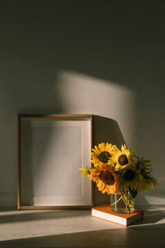 Vase of sunflowers on a hardback book next to a blank picture frame in the sunlight