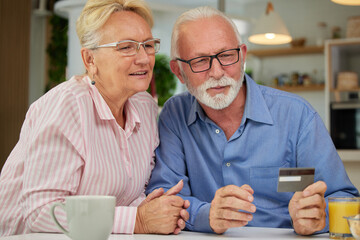 Excited and happy elderly couple shopping online from home using computer, smartphone and credit card. Modern technologies and retirement concept.