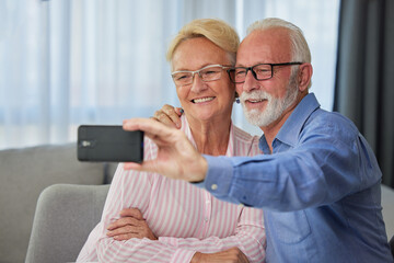 Happy senior couple taking selfie, having fun with phone camera or recording video for vlog. Smiling aged wife and husband hugging, looking at camera, posing for photo