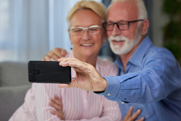 Happy senior couple taking selfie, having fun with phone camera or recording video for vlog. Smiling aged wife and husband hugging, looking at camera, posing for photo