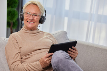 Happy senior woman wearing headphones, using tablet for social media, listening to music, video call, podcast, e-book or online job sitting on sofa at home. Modern technologies and retirement concept. - 602110488