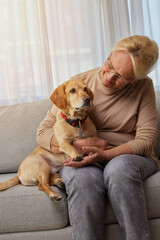 Happy retired senior woman embracing her small red dog and enjoying time with pet sitting on sofa at home