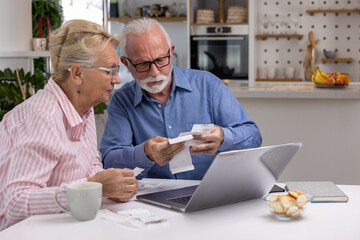 Worried senior couple checking their bills and receipts sitting at home with laptop. Frustrated elderly husband and wife having problems with finances or struggling with paying the bills online