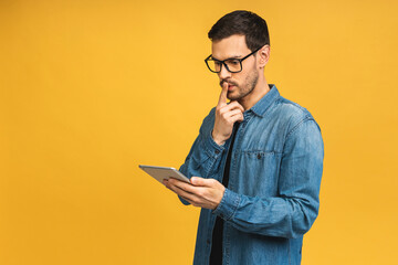 Serious thinking bearded man using digital tablet, reading media news, isolated over yellow background. - 602116261