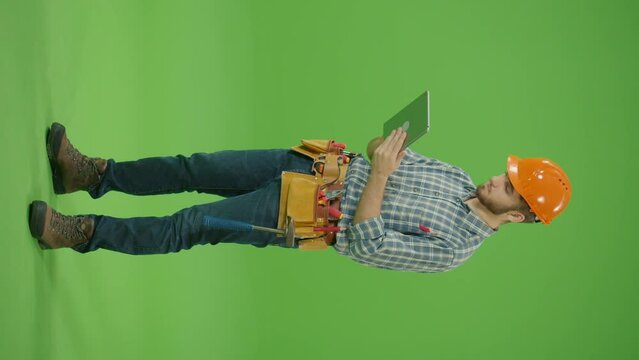 Vertical Footage.Green Screen.Side View. Young Bearded Confident Civil Engineer Wearing Checkered Shirt,Safety Helmet and Leather Tool Belt,Looking Around and Correcting the Records on the Tablet