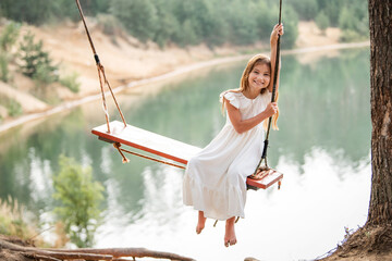 A girl in the forest swinging on a swing. Rope swing on a forest lake. Barefoot girl in a white...