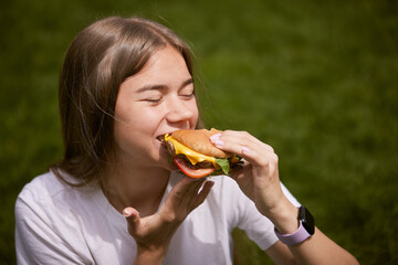 a young girl takes out a burger from a paper bag sitting on the green grass, the concept of food delivery, street food