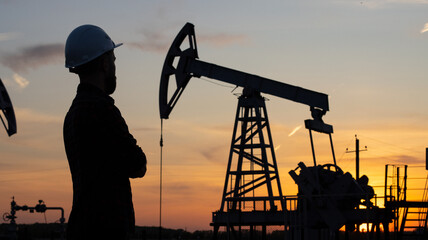 Silhouette of a young man in a construction helmet against the background of a working oil pump. An...