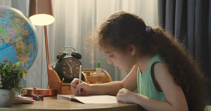 Cute smart primary school child girl learning writing doing math homework sit at home table, adorable pretty little preschool kid studying alone making notes, children elementary education concept