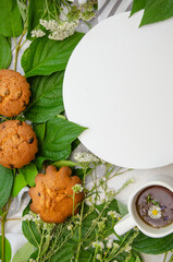 Muffins and cup of herbal tea with white round podium, wooden plate and season herbs on cotton tablecloth.card. Template for recipes or food menu
