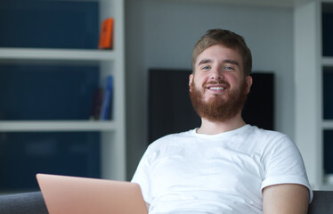Young happy positive man use laptop computer at home in living room sit on couch looking at camera and smile. Freelancer guy, freelance distant job